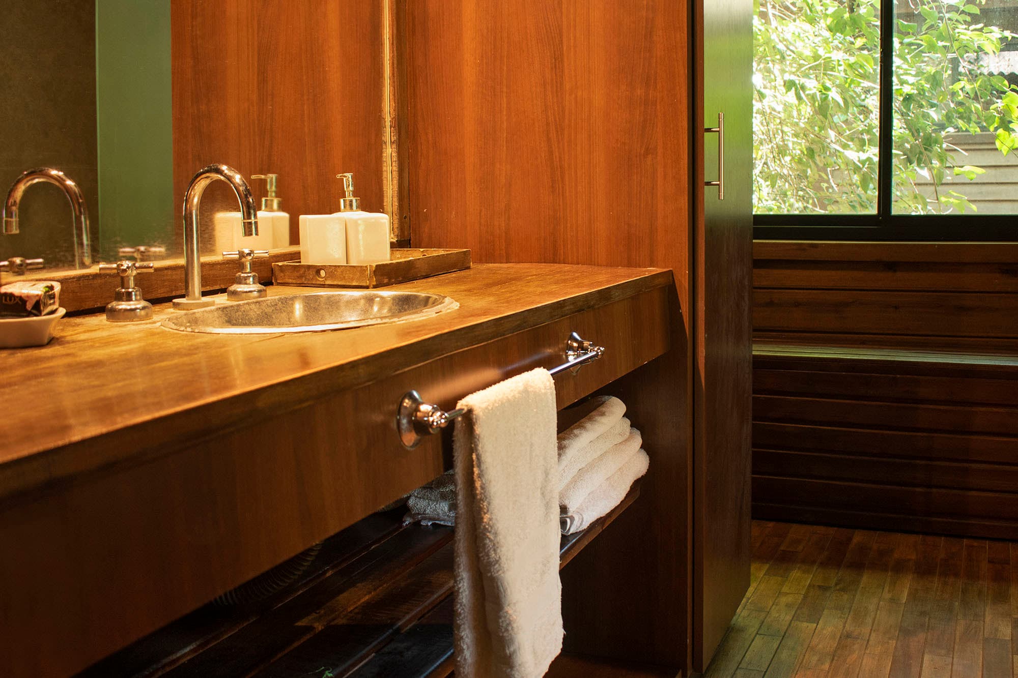 Baño de la cabaña revestido en madera, con espejo, mesada de madera oscura y bacha ovalada de metal; sobre una bandeja, los jabones y el dispenser.