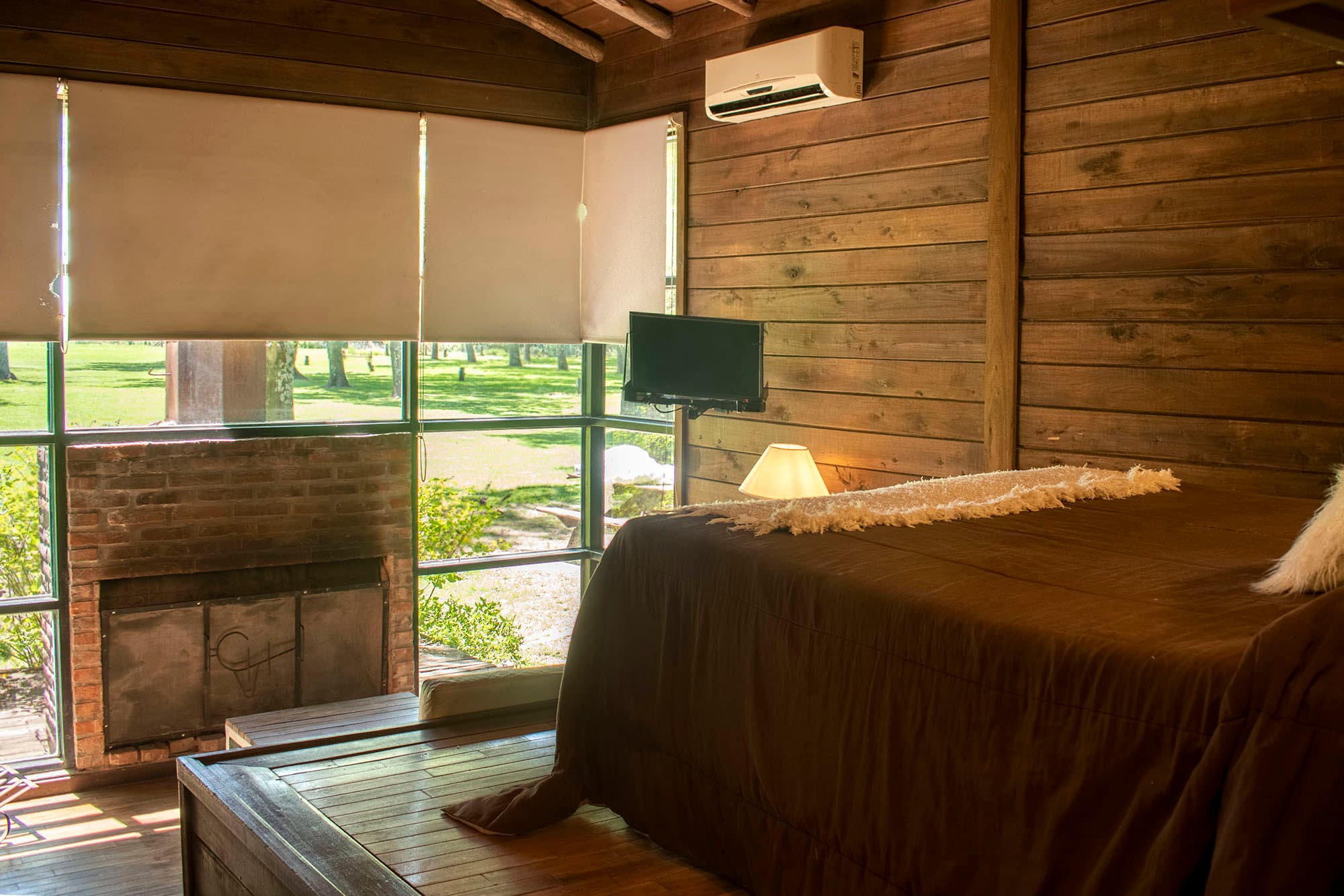 Dormitorio de una cabaña con paredes y techo de madera, cama vestida en tonos tierra, velador encendido y un ventanal esquinero que da al parque; a la izquierda, el hogar de ladrillo con la pantalla de hierro del monograma CV.