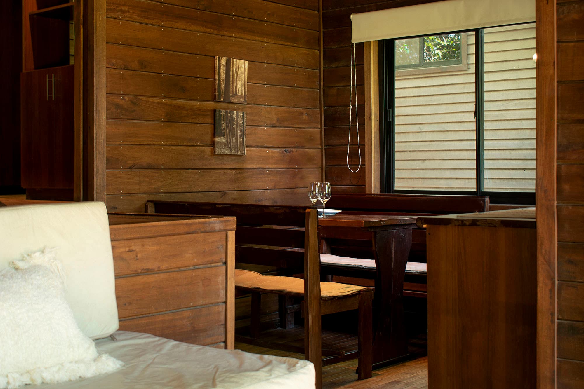 Interior de cabaña de madera con la cama en primer plano fuera de foco y, más atrás, el comedor con banco, mesa y dos copas servidas junto a la ventana.
