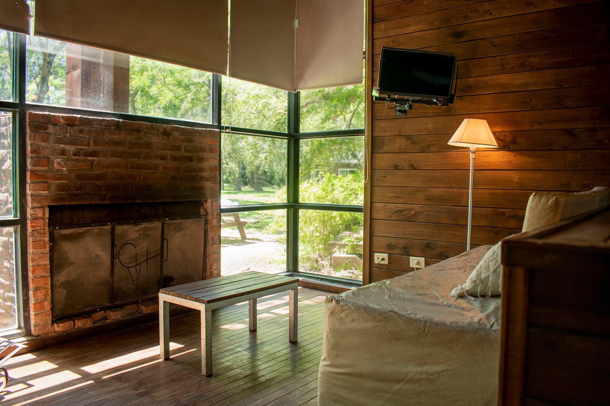 Interior de una cabaña con hogar a leña de ladrillo visto, ventanal esquinero al parque arbolado, mesa baja de madera y sillón claro.
