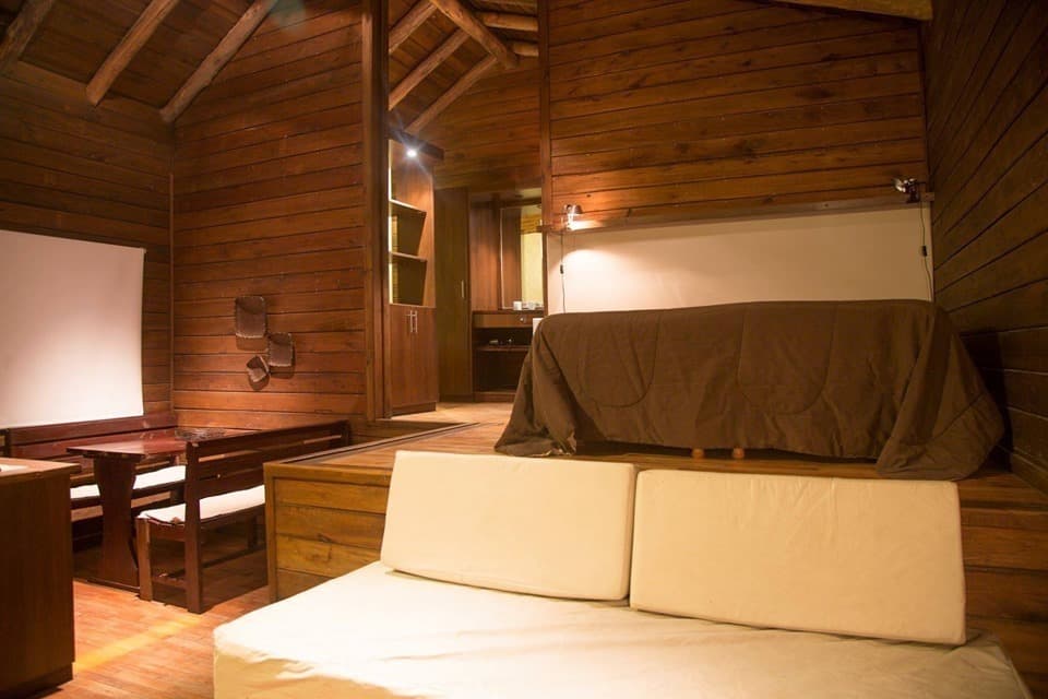 Interior de una cabaña tipo loft: la cama matrimonial sobre la plataforma alta de madera, el sofá cama abierto en el nivel de abajo y, al fondo, el paso hacia el baño.