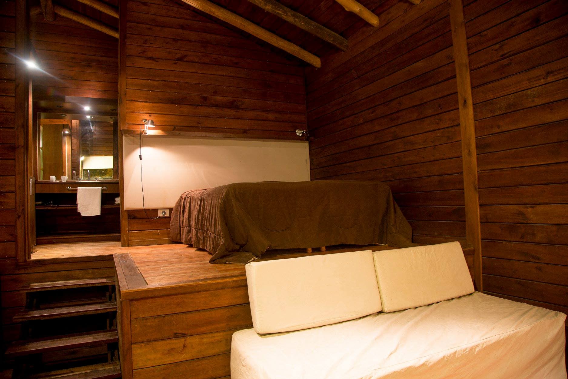 Interior de una cabaña tipo loft, con vigas de tronco en el techo: sobre una plataforma de madera, la cama con un acolchado marrón y una lámpara encendida sobre el panel claro del respaldo; a la izquierda, los escalones que bajan del nivel alto y el vano del baño, con el espejo y una toalla blanca; abajo, el sofá con funda clara.