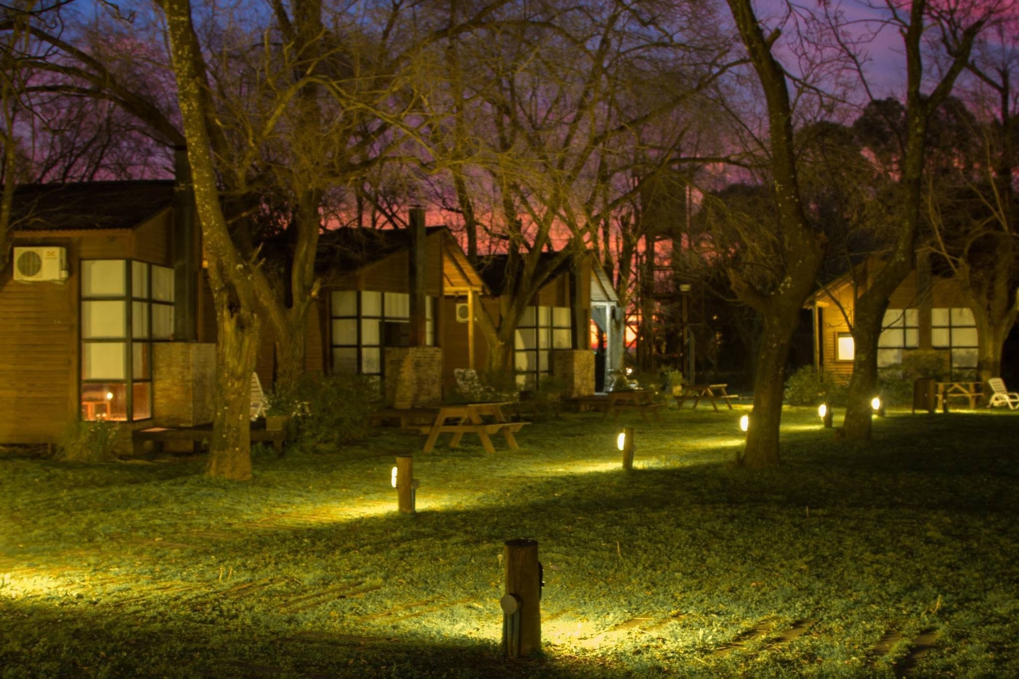 Cuatro cabañas al anochecer, con las ventanas encendidas desde adentro y el cielo todavía violeta sobre la arboleda sin hojas; en el pasto, las luces bajas del parque marcan la senda entre las mesas de picnic.