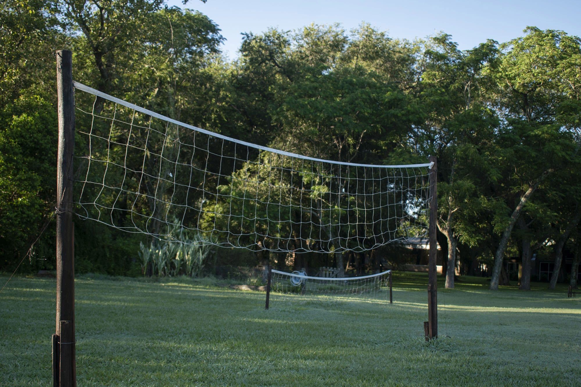 La cancha de fútbol-tenis: dos redes tendidas entre postes de madera sobre el césped, con la arboleda detrás.