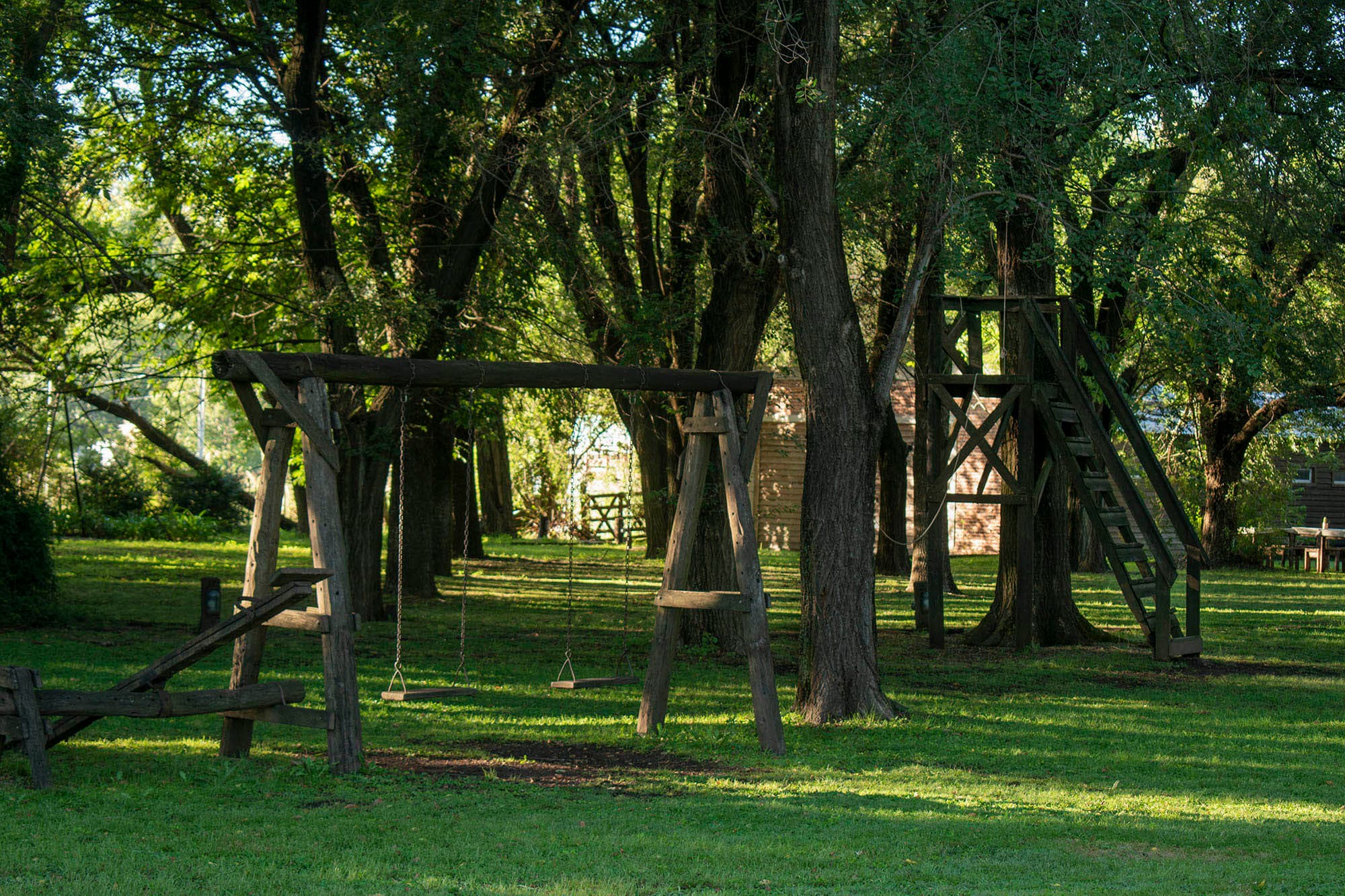 Sector de juegos sobre el pasto: hamacas dobles de madera, un sube y baja y una torre con casita, rodeados de árboles.