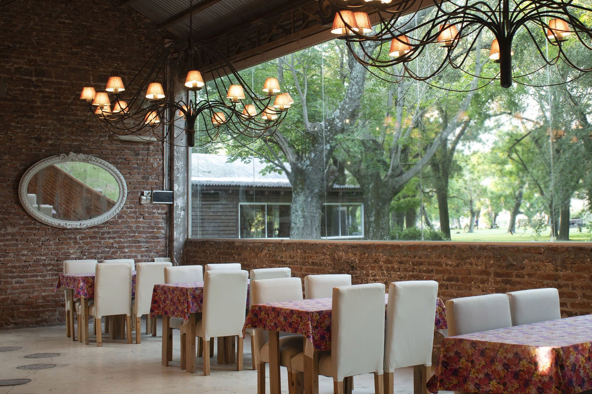 El comedor de día: paredes de ladrillo visto, dos arañas de hierro con pantallas de tela y un ventanal corrido que da al parque y a los árboles añosos; las mesas, con manteles floreados y sillas de respaldo alto.