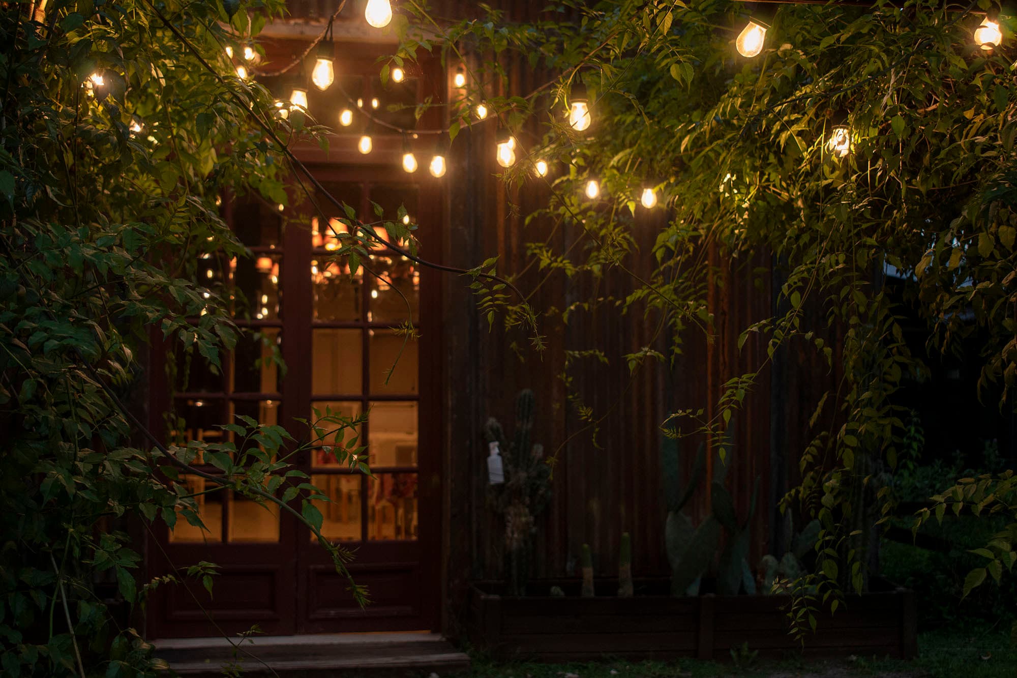 Entrada del quincho al anochecer: guirnaldas de luces cálidas entre el follaje y la puerta vidriada con el comedor iluminado del otro lado.