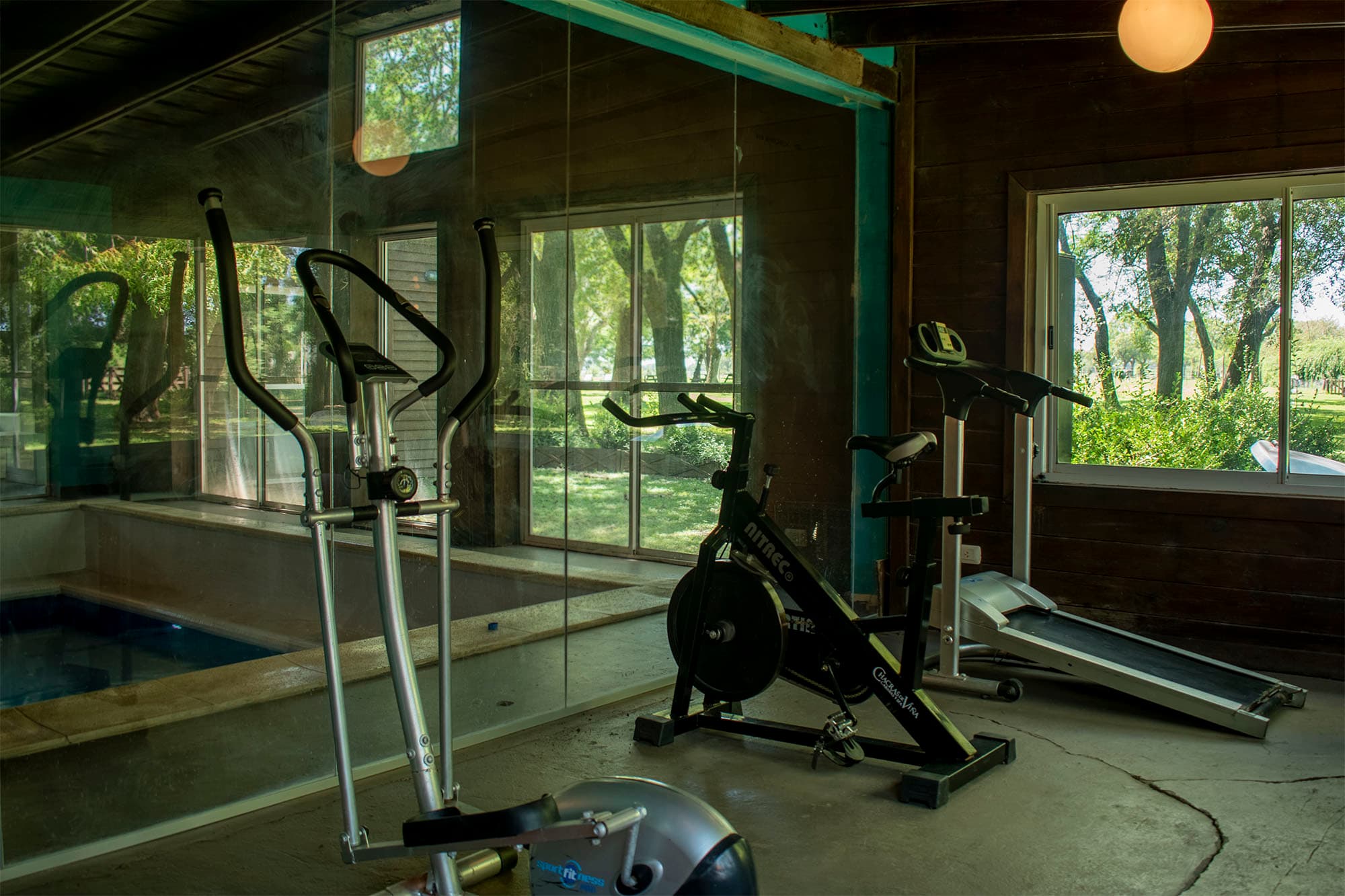 Mini gimnasio del spa: elíptico, bicicleta fija y cinta frente a los ventanales, con la pileta cubierta del otro lado del vidrio.