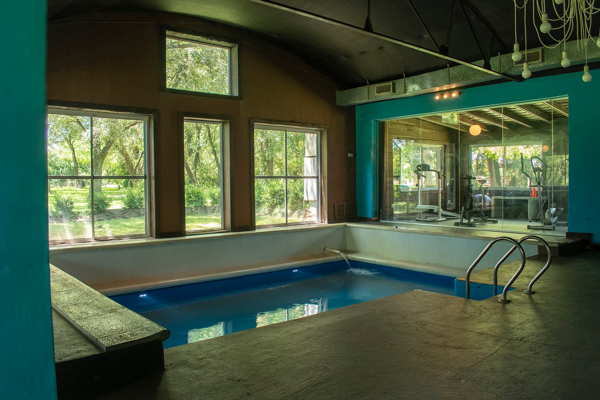 Vista general de la pileta cubierta: el agua azul, los ventanales altos al parque y el mini gimnasio detrás del vidrio, bajo el techo abovedado.