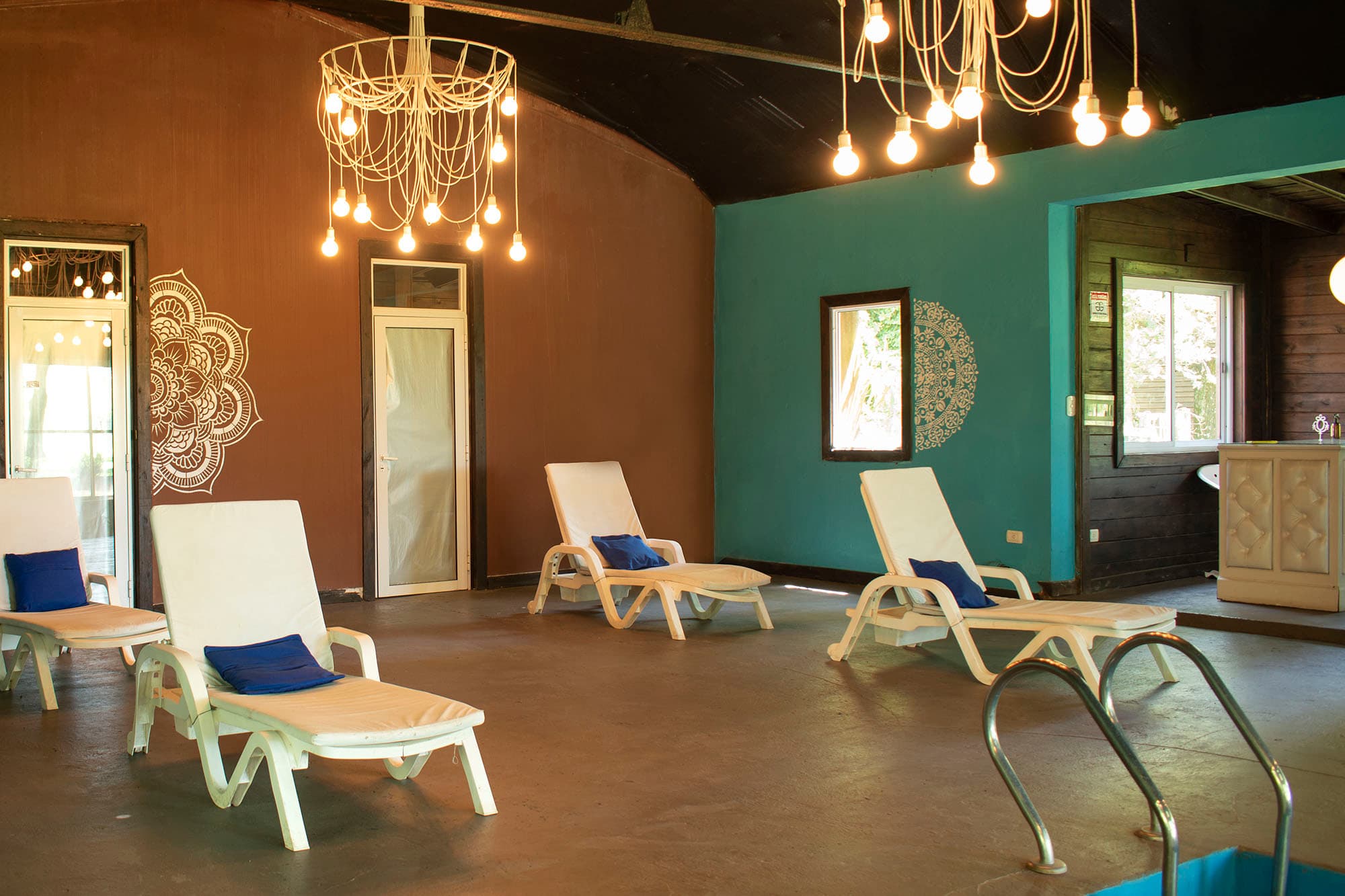 Sector de relax junto a la pileta cubierta: reposeras blancas con almohadones azules contra los muros terracota y verde, bajo dos lámparas colgantes.